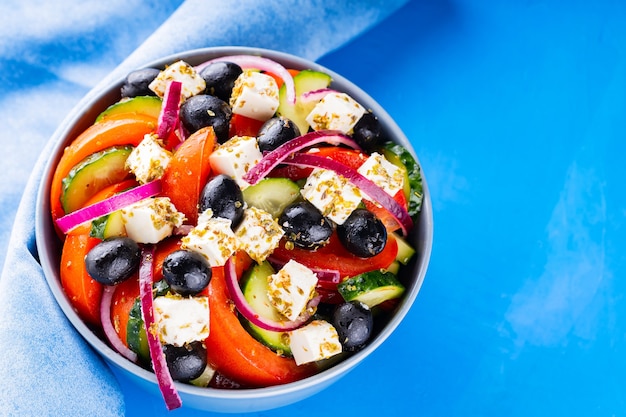 青い背景にギリシャ風サラダと青いナプキン。新鮮な野菜、フェタチーズ、ブラックオリーブ。ギリシャ料理。上面図。コピースペース