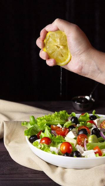 Greek salad in a plate on a dark background. Healthy vegetarian food. The hand squeezes out the lemon juice. Feta cheese, lettuce leaves, cherry tomatoes.