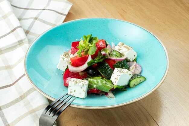 Greek salad with olives, tomatoes, feta cheese, onions in blue bowl on wooden wall. Close up. Selective focus