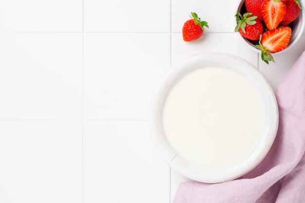 Greek yogurt in white bowl with ingredients for making breakfast granola and fresh strawberries on white table