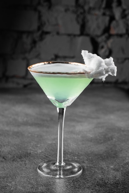 Green alcohol cocktail in a glass decorated with cotton candy on gray surface.