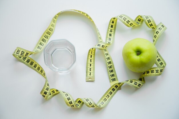 A green apple and a glass of water stand on a white background they are entwined with a measuring ta...