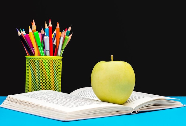 Green apple on opened book with colored pencils against chalkboard Back to school