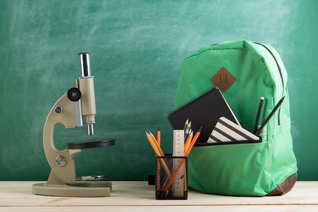 Зеленый рюкзак, микроскоп, черные тетради и карандаши на фоне доски