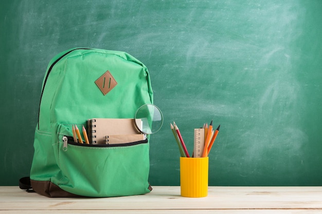 Green backpack notebooks and school supplies on the background of the blackboard