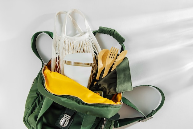 Zaino verde con borsa da mercato in rete, posate in bambù e tazza da caffè riutilizzabile.