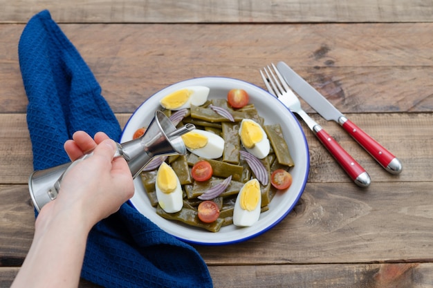 Green bean salad with boiled egg and tomato. Woman's hand with oilcan. Copy space. Wooden background.