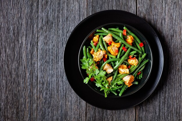 Green beans with curried salmon fish and bell peppers served on a black plate with parsley, lemon wedge, on a dark wooden surface with golden cutlery, top view, flat lay, close-up