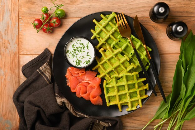 Green Belgian waffles Spinach or wild garlic or pesto waffles with red salmon and cream sauce on black plate over dark rustic wooden background Delicious breakfast snack brunch Top view Mock up