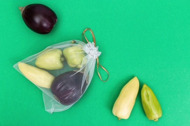 Green bell peppers and eggplants in reusable eco bags on green paper background. Fresh vegetables in bags from transparent textile for food storage. Plastic free concept. Flat lay. Top View.