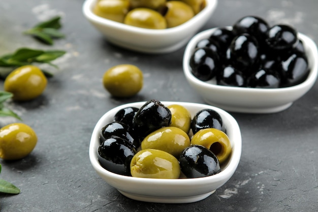 Green and black olives in a white ceramic bowl with leaves on a dark graphite background. close-up