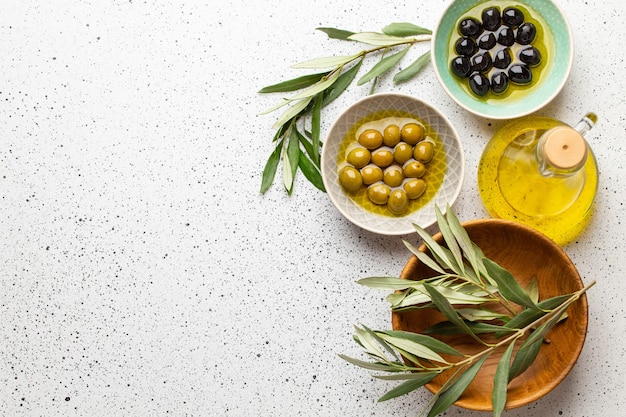 Green and black olives with olive oil in a glass bottle, olive tree sprigs and cut fresh ciabatta bread on wooden cutting board. White rustic background, healthy mediterranean food, space for text