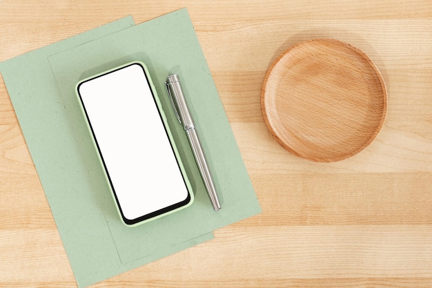 Green blank smart phone on wooden office table Recycled paper Business paper pen and cell phone Mock Up