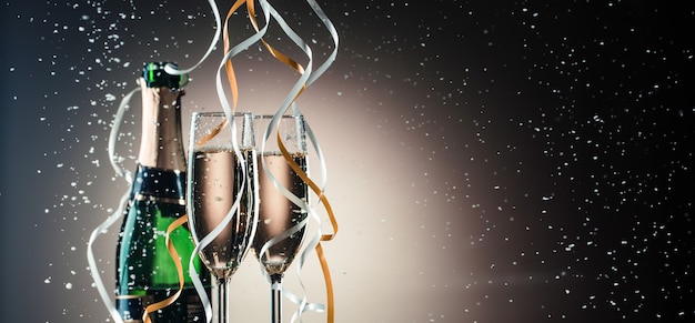 Green bottle of champagne, wine glasses with silver and golden ribbons close-up. Festive composition on a dark background with backlight