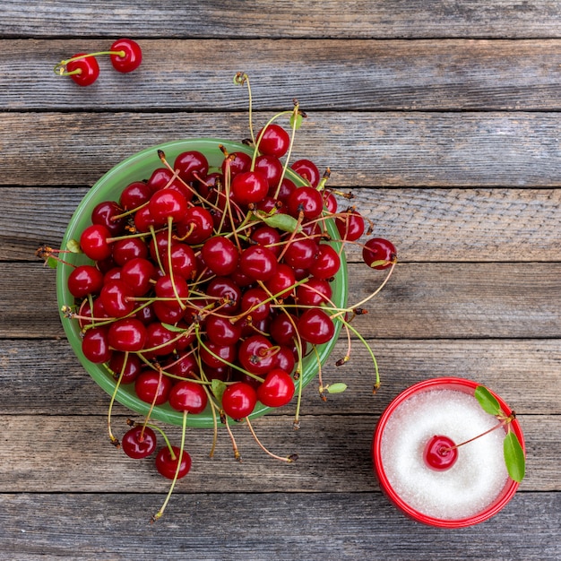 Green bowl with ripe cherry berries and cup with sugar on a wooden background, close up top view