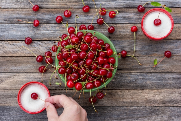 green bowl with ripe cherry berries and a male hand dips a cherry berry in a cup with sugar on a wooden table, closeup top view