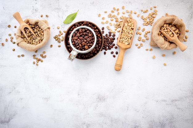 Green and brown decaf unroasted and dark roasted coffee beans in hemp sack bags with scoops setup on white concrete.