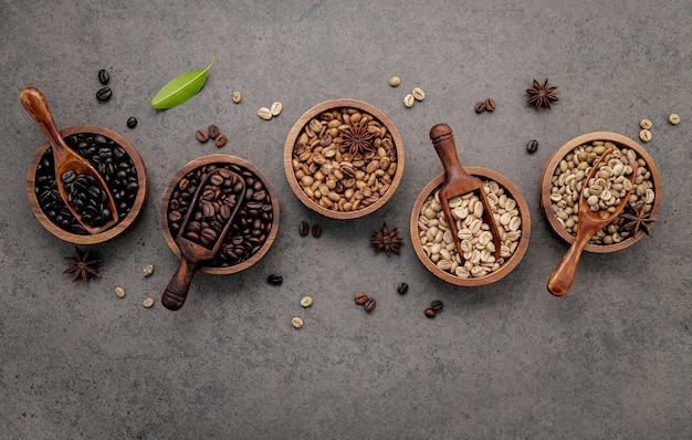 Green and brown unroasted and dark roasted coffee beans in wooden bowl with spoons set up on dark concrete.