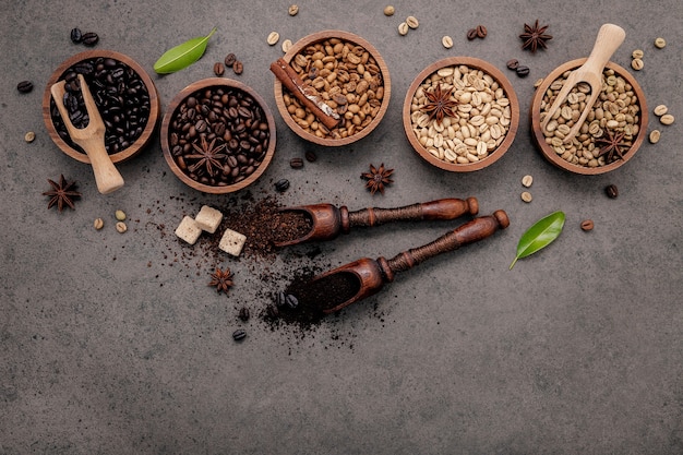 Green and brown unroasted and dark roasted coffee beans in wooden bowl with spoons set up on dark concrete.