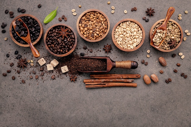 Green and brown unroasted and dark roasted coffee beans in wooden bowl with spoons set up on dark concrete.