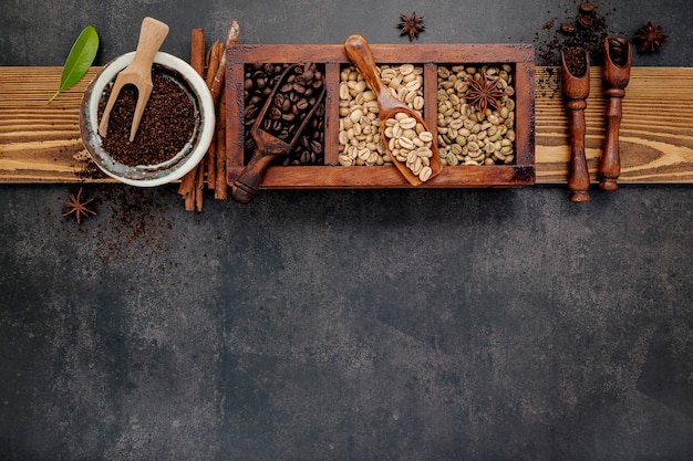 Green and brown unroasted and dark roasted coffee beans in wooden box with scoops setup on dark stone.