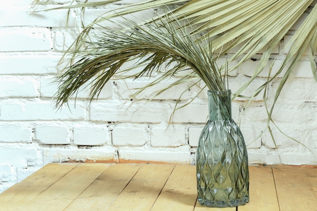 Photo green dry branch of home date palm in beautiful glass bottle of blue glass on background of white brick wall.