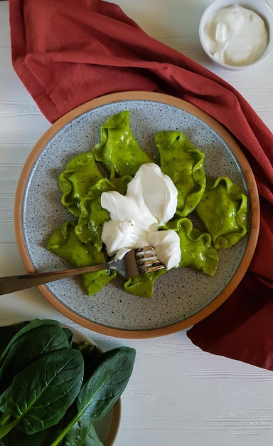 Green dumplings with cottage cheese, spinach and sour cream on ceramic plate with red textiles on white background. vertical, top view