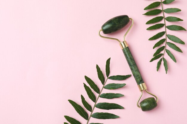 Green face roller and leaves on pink background