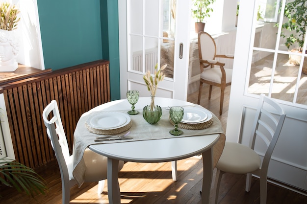 Green glasses and white dishes on the kitchen table, with green plant on living room in the background.