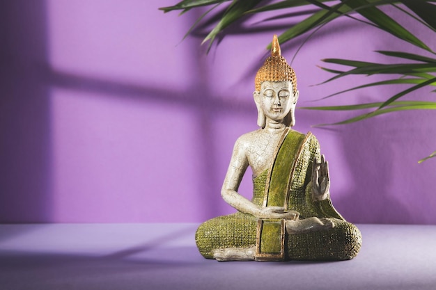 Green and golden buddha statue in meditation on light purple\
background with copy space