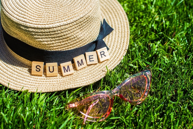 緑の芝生の上に麦わら帽子、サングラス、木製の文字。夏という言葉。夏の背景。休暇、週末。