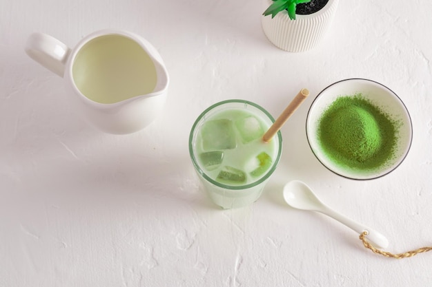 Зеленый полезный чай матча латте с кубиками льда на белом столе с миской порошка матча и молочным кувшином с молочными аминокислотами, вид сверху