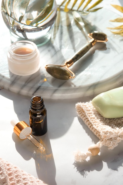 Green jade face roller massager and face care products on\
marble stone background glass crystal ball and exotic palm leaves\
towel soap skincare tonic essential oil serum