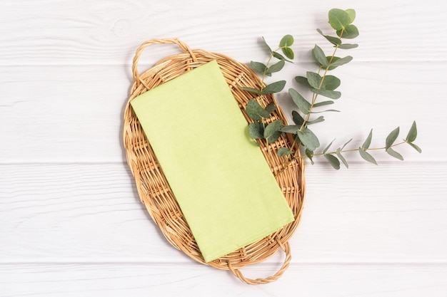 Green kitchen tea towel and eucalyptus branch on a wicker tray on a white wood table