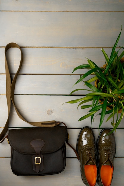 Green lacquered oxford shoes and crossbody bag on wooden background near flower pot. 