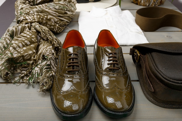 Photo green lacquered oxford shoes near white shirt, bag and scarf on wooden background. back view.