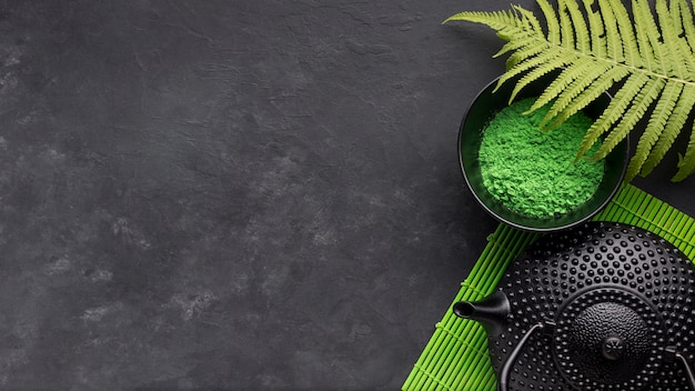 Green match tea powder and fern leaves with black teapot on black background