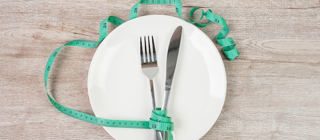 Green Measuring tape wrapped around fork and knife with white ceramic dish on wooden table background