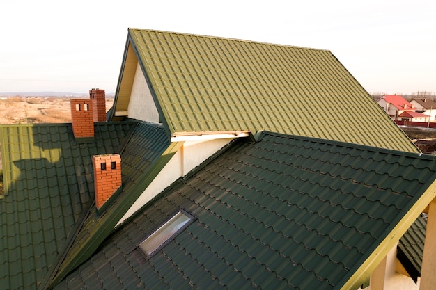 Photo green metal shingled house roof with attic plastic window and brick chimney.