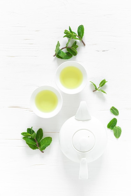 Green mint tea with fresh leaves in cups and teapot overhead on white wooden table healthy warming drink antioxidant beverage top view