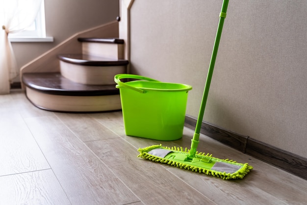 Green mop standing near white wall. Cleaning gear. Green plastic bucket. Householding.