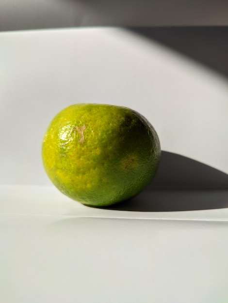 A green orange on a white surface with a shadow on it.