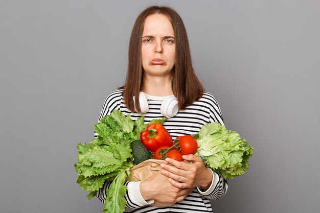 最適な健康のためのグリーンとオーガニック 灰色の背景に野菜を持った悲しい動揺の女性は、健康的な食事の代わりにファーストフードを望んでいます