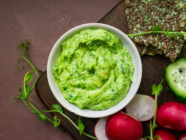 Photo green pea hummus, fresh greens, vegetables, ckackers served on a wooden board, closeup view