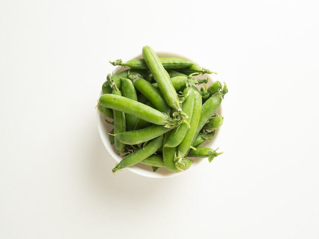 Green pea pods in a white bowl on a white table, top view