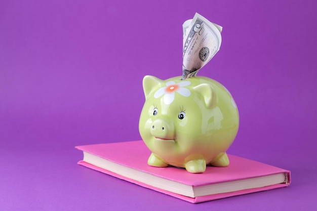 Зеленая свинья копилка и деньги и книга на ярко-фиолетовом фоне. Финансы, сбережения, деньги. место для текста