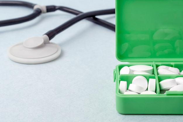 テーブルの上の白い錠剤と緑のピルボックス。背景には、聴診器。