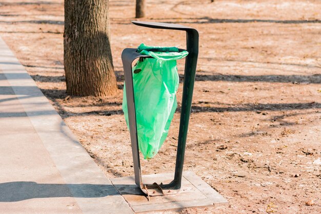 Green plastic bag hung to the metallic construction in the\
public park. trash bag. rubbish can. bin. outdoor. pathway. iron.\
city. urban