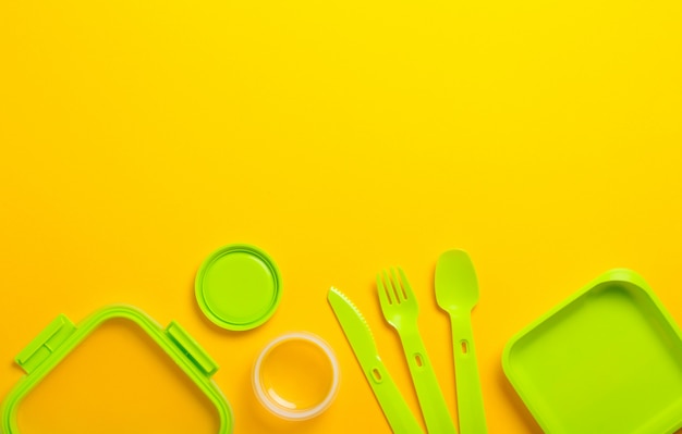 黄色の背景にフォーク、スプーン、ナイフで緑のプラスチック製ランチボックス。平面図、フラットが横たわっていた。学校やオフィスの食品容器。コピースペース。