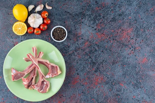 Green plate of uncooked lamb limbs with vegetables on marble.